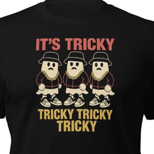 May include: Black t-shirt featuring a graphic with the text "It's Tricky" above three cartoon figures wearing hats and collars. The phrase "Tricky Tricky Tricky" is printed below the figures in a gold color.