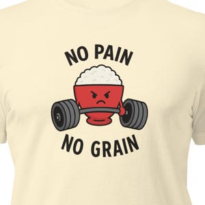 Peut inclure: T-shirt crème avec un dessin animé d'un bol de riz soulevant une barre. Le bol de riz a une expression de colère. Le texte "NO PAIN NO GRAIN" est imprimé au-dessus et en dessous du dessin en noir.