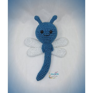 May include: A blue crocheted dragonfly stuffed animal with white wings. The dragonfly has black eyes and a smiling mouth.