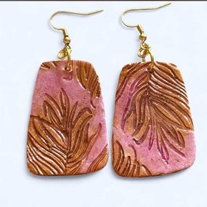 May include: A pair of dangle earrings featuring rectangular, slightly tapered pendants with a textured, metallic gold palm leaf pattern embossed onto a shimmering pink background. The earrings are suspended from gold-toned metal fishhook wires.