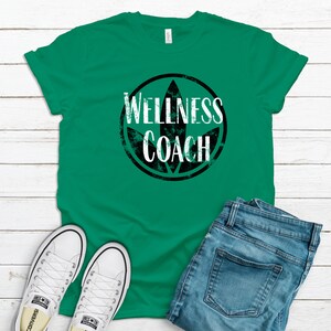 May include: A green t-shirt with a black circle and white text that says "Wellness Coach". The circle has a green leaf design inside. The t-shirt is laid out on a white wooden surface with a pair of white sneakers and blue jeans.