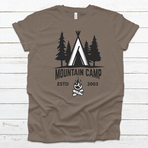 Könnte beinhalten: Ein braunes T-Shirt mit einem schwarz-weißen Grafikdesign eines Zeltes, Bäumen und einem Lagerfeuer. Der Text "Mountain Camp" ist in einem schwarzen Banner über dem Lagerfeuer geschrieben. Der Text "Estd 2003" ist unter dem Lagerfeuer geschrieben.