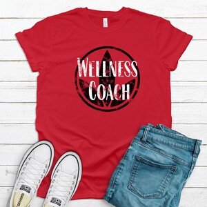 May include: Red t-shirt with a black and white distressed circle graphic that says "Wellness Coach". The graphic features a stylized leaf design. The shirt is laid flat on a white wood surface with a pair of white sneakers and blue jeans.