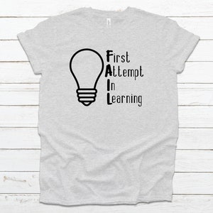 May include: Light gray t-shirt with the text "FAIL" in a vertical arrangement, with a lightbulb graphic above the letters. The text "First Attempt In Learning" is printed in black. The shirt is made of a soft material.