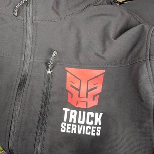 Pu&ograve; includere: Giacca softshell nera con un logo rosso di un robot e il testo "TRUCK SERVICES" stampato sul davanti.