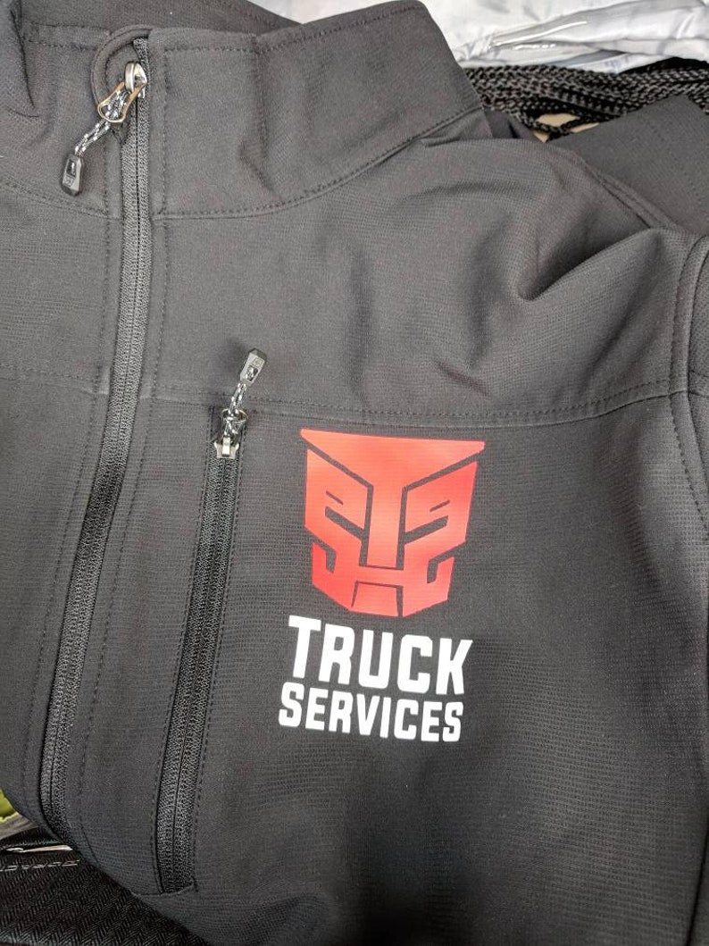 Pu&ograve; includere: Giacca softshell nera con un logo rosso di un robot e il testo "TRUCK SERVICES" stampato sul davanti.
