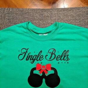 May include: Green t-shirt with black text that says "Jingle Bells" and a red bow on top of two black kettlebells.