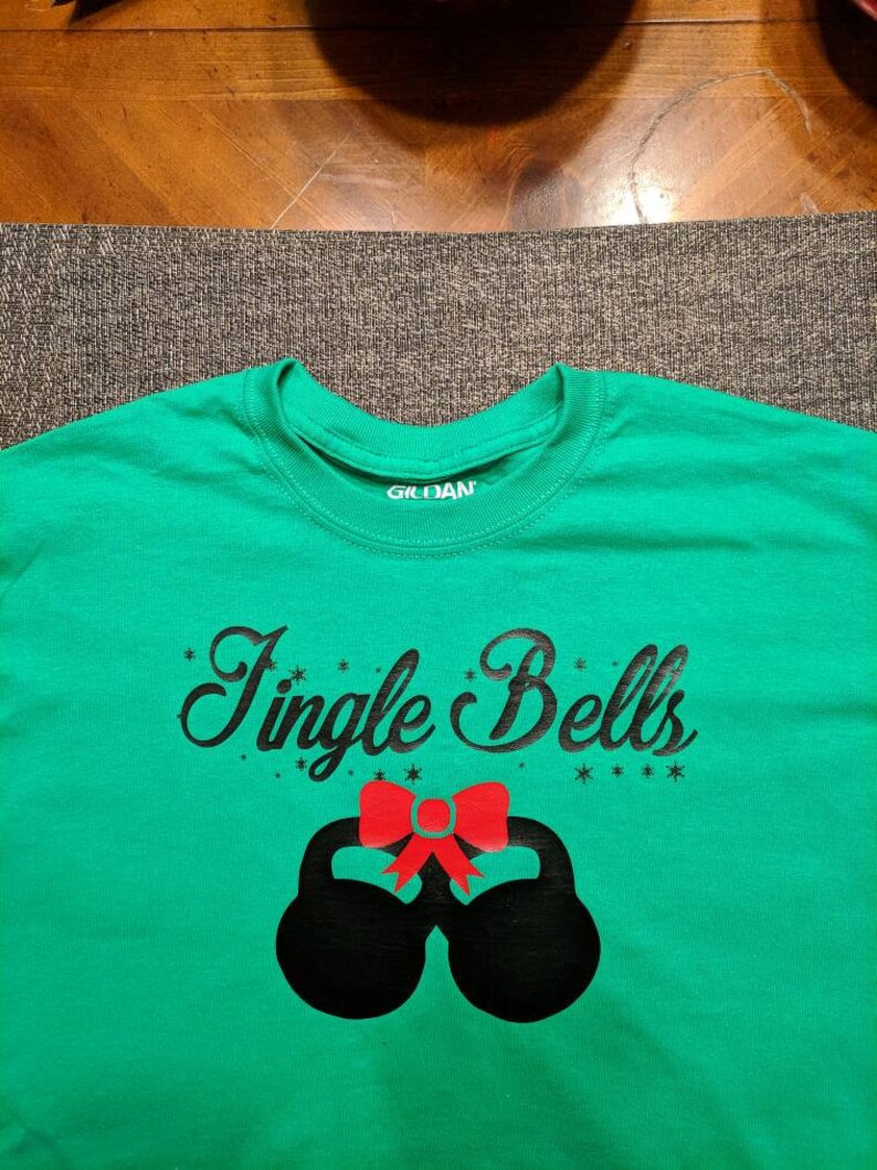 May include: Green t-shirt with black text that says "Jingle Bells" and a red bow on top of two black kettlebells.