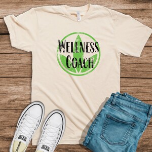 May include: A beige t-shirt with a black and green graphic that says "Wellness Coach".