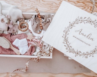 May include: A wooden box filled with baby items, including a knitted sweater, socks, a rattle, and a stuffed mouse. The lid of the box has a floral design and the text "Amelia 28.06.2021 3250g | 04:32 Uhr".