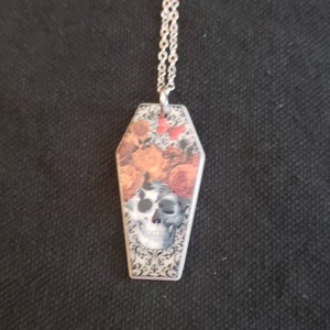 May include: A silver chain necklace with a coffin-shaped pendant. The pendant features a skull with floral accents and a black and white patterned background.