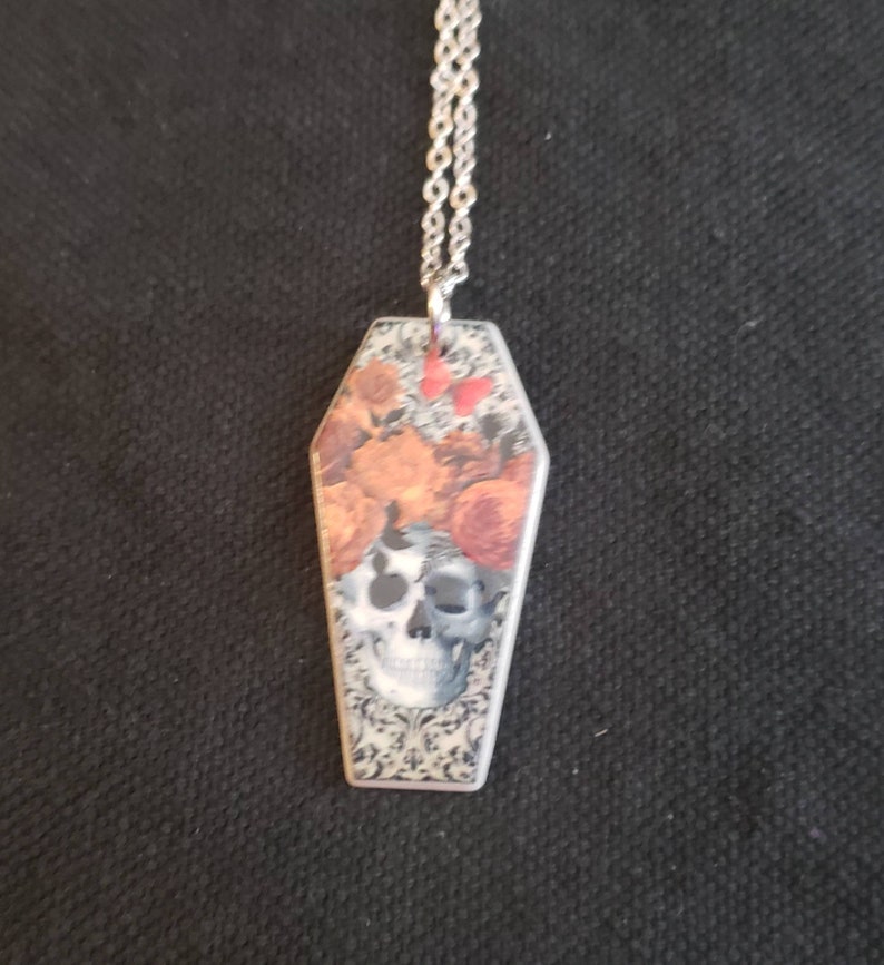May include: A silver chain necklace with a coffin-shaped pendant. The pendant features a skull with floral accents and a black and white patterned background.