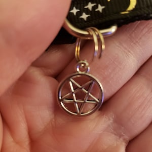 Puede incluir: Un collar de patr&oacute;n blanco y negro con una luna creciente dorada y estrellas blancas. Un amuleto plateado con un pentagrama est&aacute; unido al collar.