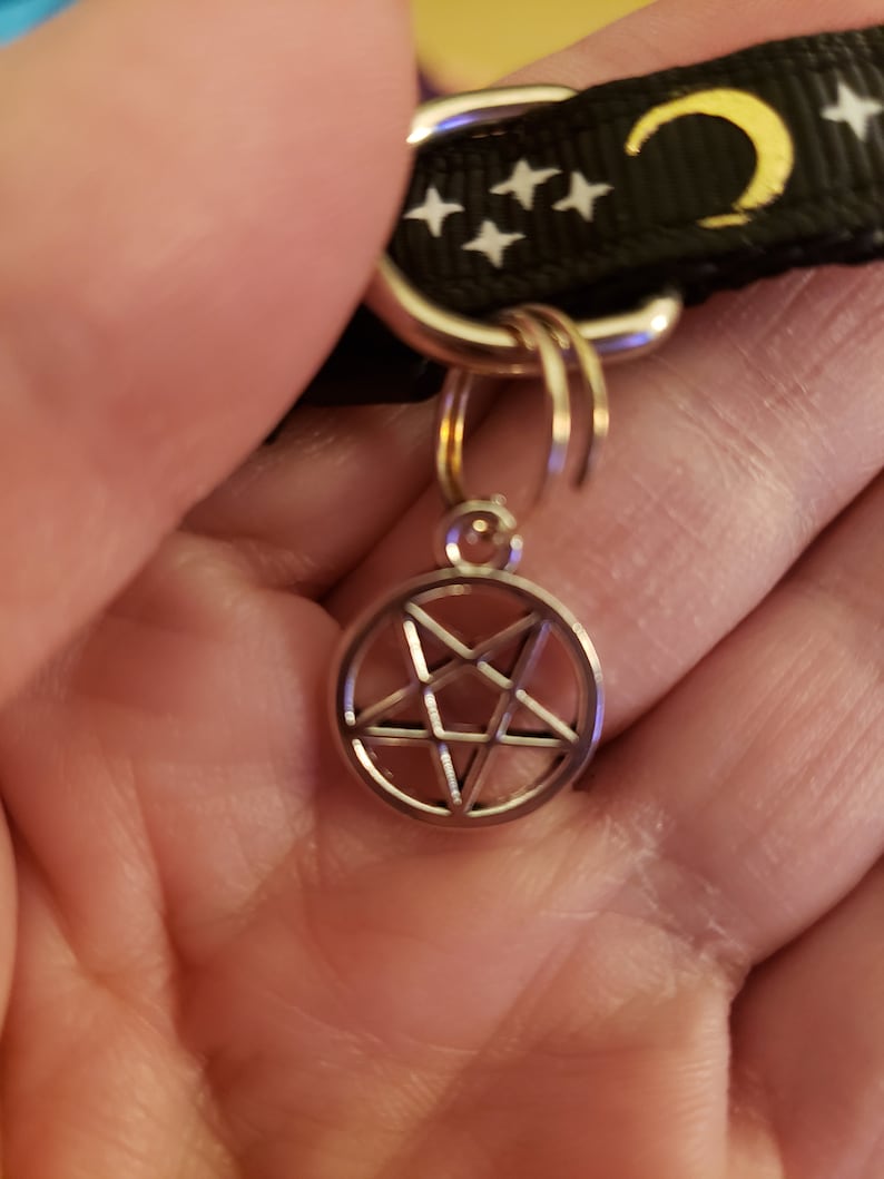 Puede incluir: Un collar de patr&oacute;n blanco y negro con una luna creciente dorada y estrellas blancas. Un amuleto plateado con un pentagrama est&aacute; unido al collar.