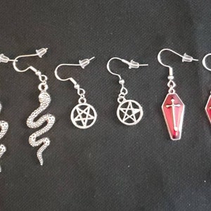 May include: Six pairs of silver earrings with different charms. Two pairs feature a silver snake design, two pairs feature a pentagram, and two pairs feature a red coffin with a silver cross.