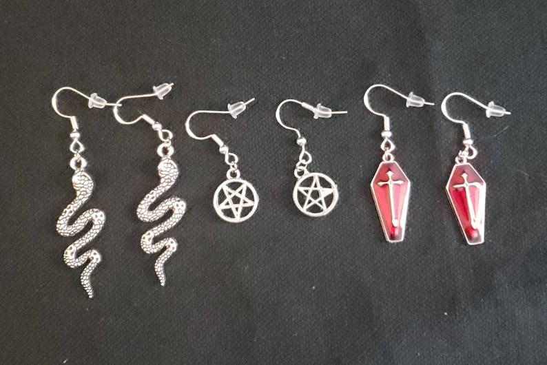 May include: Six pairs of silver earrings with different charms. Two pairs feature a silver snake design, two pairs feature a pentagram, and two pairs feature a red coffin with a silver cross.