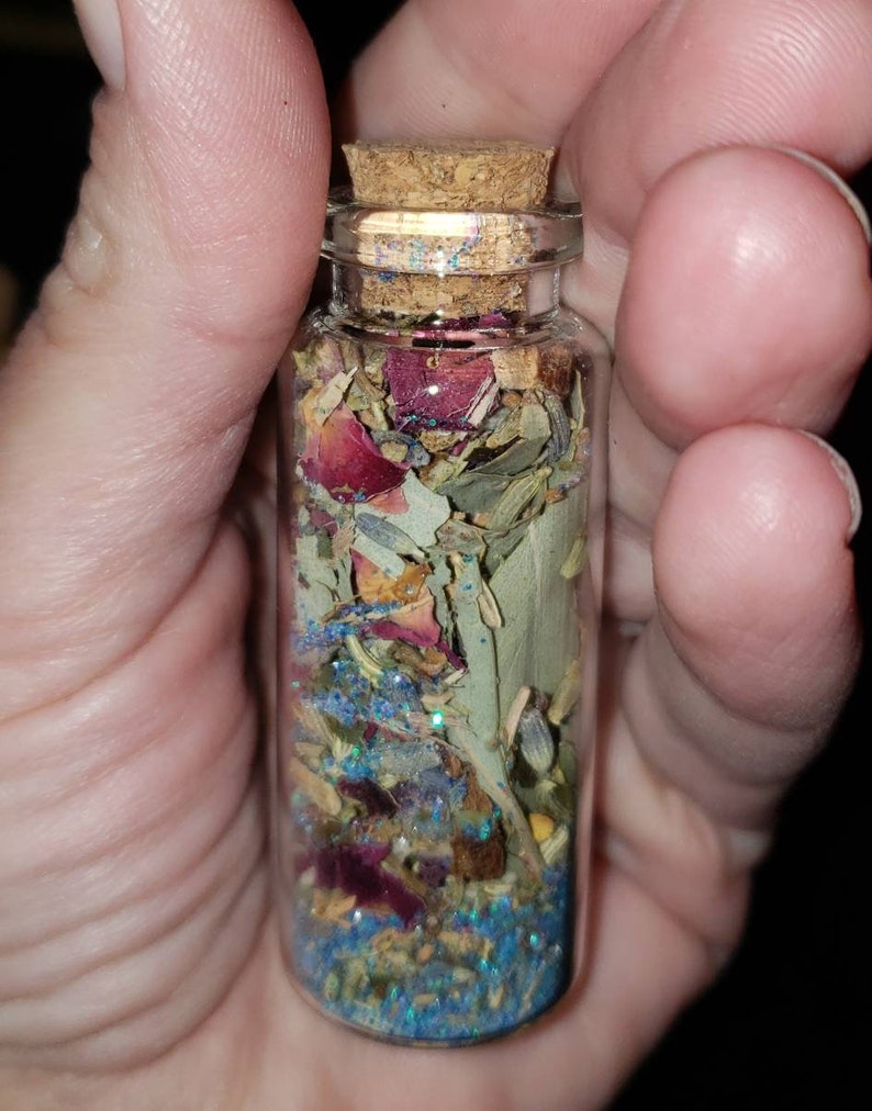 May include: A small glass bottle with a cork stopper filled with dried herbs, flowers, and blue glitter.