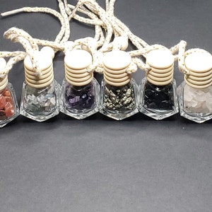 May include: Seven small glass bottles with wooden tops and string hangers filled with various colored crystals. The bottles are arranged in a row on a black surface.