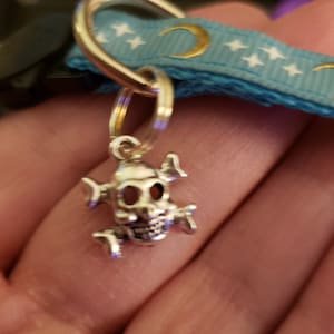 Puede incluir: Encanto de calavera de plata con huesos cruzados, unido a un llavero dorado. El encanto cuelga de una cinta azul con medias lunas doradas y estrellas blancas.