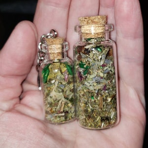 May include: Two glass bottles with cork tops filled with dried herbs and green glitter. The bottles are different sizes and are held in a hand.