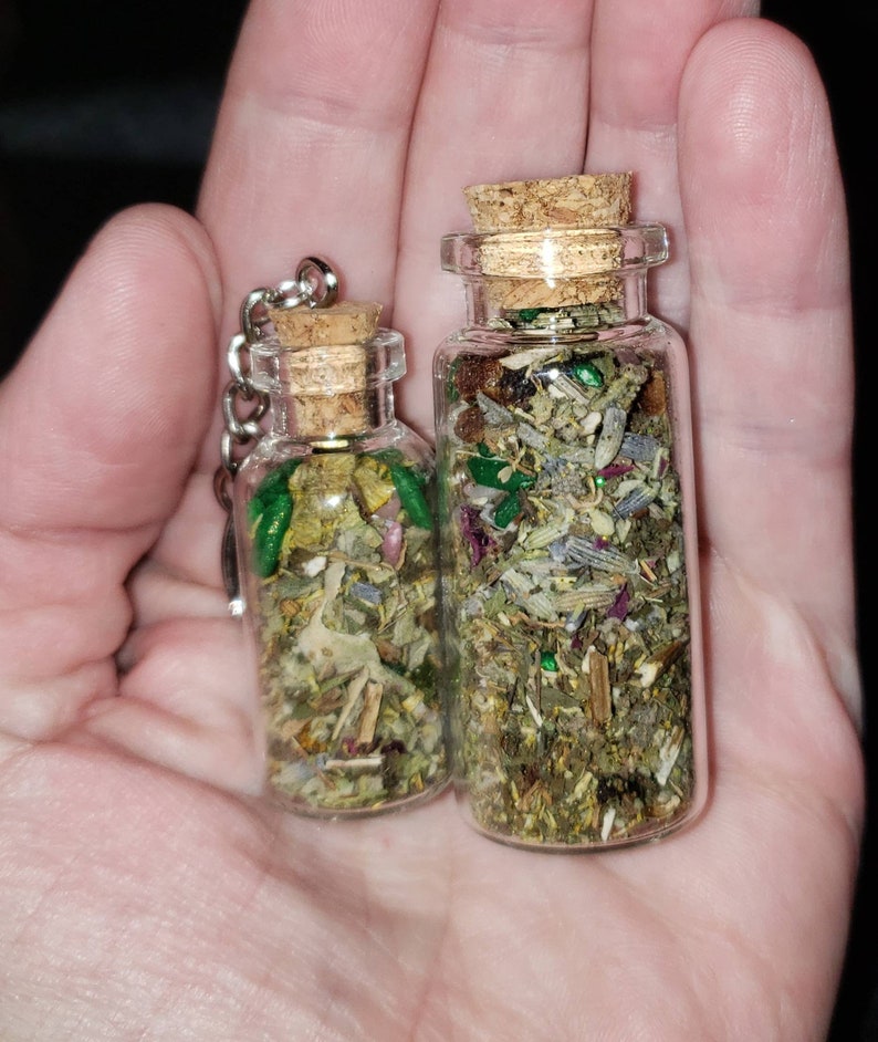 May include: Two glass bottles with cork tops filled with dried herbs and green glitter. The bottles are different sizes and are held in a hand.