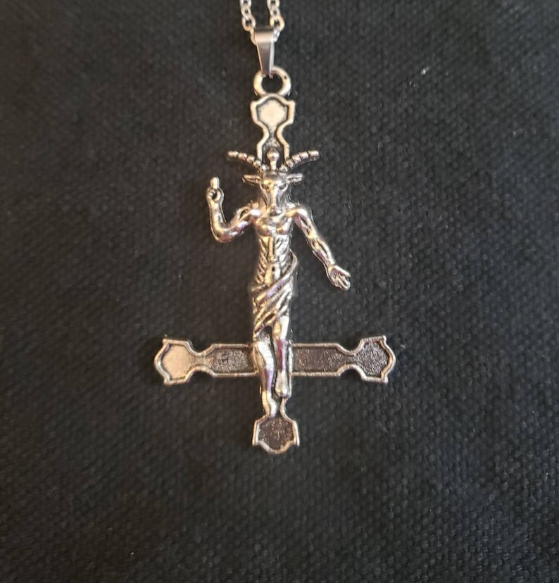 May include: Silver pendant necklace featuring a depiction of a goat-headed figure on an inverted cross.