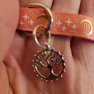 Puede incluir: Un amuleto de plata con un dise&ntilde;o de &aacute;rbol cuelga de una cinta rosa con estrellas blancas y medias lunas doradas.