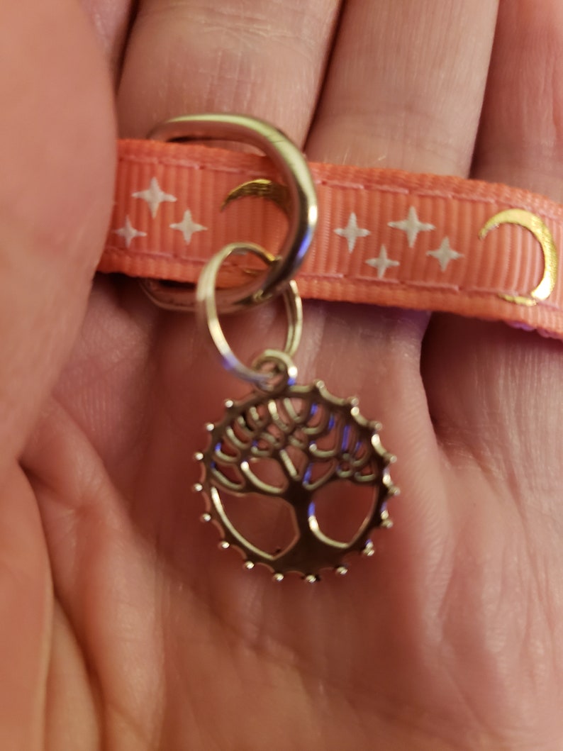 Puede incluir: Un amuleto de plata con un dise&ntilde;o de &aacute;rbol cuelga de una cinta rosa con estrellas blancas y medias lunas doradas.