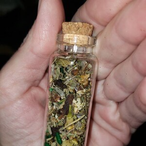 May include: A small glass bottle with a cork stopper filled with dried herbs and green glitter. The bottle is held in a hand.