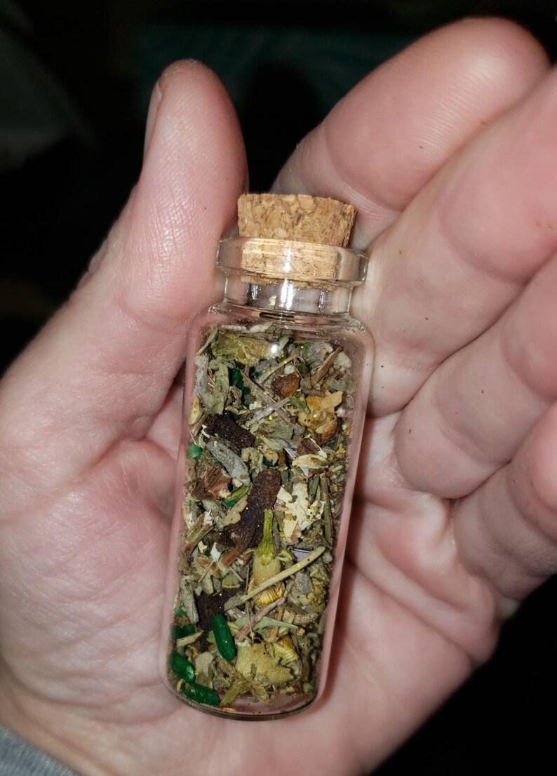 May include: A small glass bottle with a cork stopper filled with dried herbs and green glitter. The bottle is held in a hand.