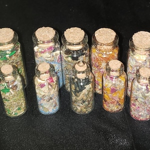 May include: Ten small glass bottles with cork tops filled with various dried herbs, flowers, and other natural materials. The bottles are arranged in two rows on a black surface.
