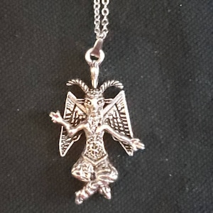 May include: Silver pendant necklace featuring a detailed depiction of a winged demon with horns and a goatee. The pendant is hanging from a silver chain.