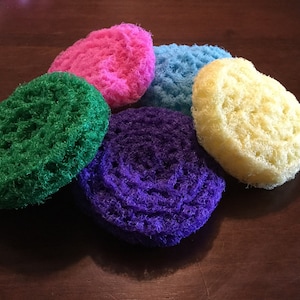 May include: Five colorful, round, crocheted scrubbers in green, purple, pink, blue, and yellow.