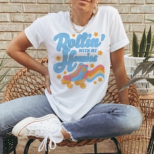 May include: White t-shirt with a graphic of a roller skate and the text "Rollin' with my Homies". The person wearing the shirt is sitting on a wicker chair.