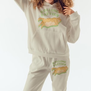 May include: A cream-colored hoodie and sweatpants set with a graphic of a cartoon orange cat on each piece. The text "Rolling Fatties" is printed in green on both the hoodie and sweatpants.