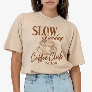 May include: A beige t-shirt with a brown graphic that says "Slow Sunday Coffee Club Est. 1990" and a drawing of a coffee cup with steam.