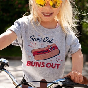 K&ouml;nnte beinhalten: Ein junges Kind tr&auml;gt ein graues T-Shirt mit einem Hotdog-Motiv und dem Text "Suns Out, Buns Out" und f&auml;hrt ein kleines Fahrrad mit einem Weidenkorb.