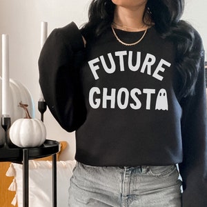 May include: A black sweatshirt with the text "FUTURE GHOST" in white lettering and a small ghost graphic. The sweatshirt is being worn by a person with long dark hair and blue jeans.