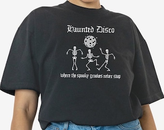 May include: Dark grey t-shirt featuring the words "Haunted Disco" above a graphic of three dancing skeletons and a disco ball. The text "Where the spooky grooves never stop" is below. Worn with blue denim shorts.