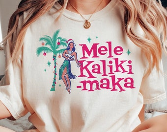 May include: Cream-colored t-shirt with a tropical Christmas design. The graphic features a woman in a Santa hat, a palm tree, and the words "Mele Kalikimaka" in pink. The shirt is part of a casual wear collection.