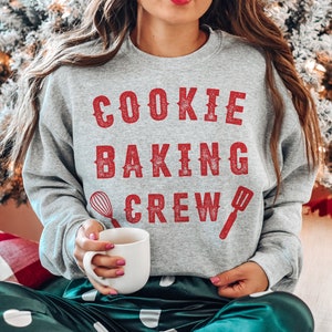 May include: A gray sweatshirt with the words "COOKIE BAKING CREW" in red, along with illustrations of a whisk and spatula. The person is holding a white mug. The person is wearing green satin pajama pants with white polka dots.