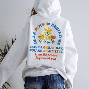 May include: White hoodie with a colorful graphic on the back that says "Dear Person Behind Me Have a Great Day, You're Beautiful from the person in front of you".