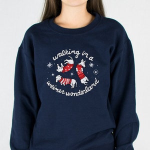 May include: Navy blue sweatshirt with a circular graphic featuring three dachshunds in red and white sweaters, surrounded by snowflakes. The text "walking in a weiner wonderland" is arched around the dogs. The sweatshirt is worn by a person.