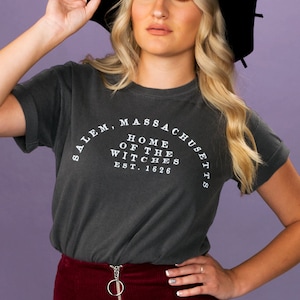 Puede incluir: Una mujer con un sombrero negro de ala ancha y una camiseta gris antracita con el texto "Salem, Massachusetts, Home of the Witches, Est. 1626" impreso en ella. Lleva una falda color burdeos.