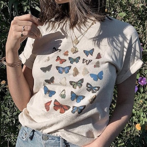 May include: A cream colored t-shirt with a graphic print of colorful butterflies. The shirt has a crew neck and short sleeves.