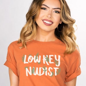 May include: An orange t-shirt with the text "LOW KEY NUDIST" in white, with green leaf accents. The shirt is worn by a person with wavy blonde hair and gold hoop earrings.
