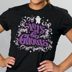 May include: Black t-shirt with the phrase "Let's Go Ghouls" in purple and white, surrounded by a decorative design of swirling smoke and stars. A small white ghost graphic is above the text. The shirt is a crew neck.