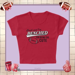 May include: Red crop top with the text "Coach said I'm Benched for Being Too Cute" and a football graphic. The shirt has a round neckline and short sleeves. The background is a light purple color with football-themed decorations.
