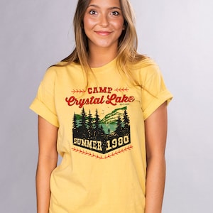 May include: Yellow t-shirt with a vintage-style graphic design that says "Camp Crystal Lake Est. 1955 Summer 1980". The graphic features a forest scene with a lake and a person fishing.