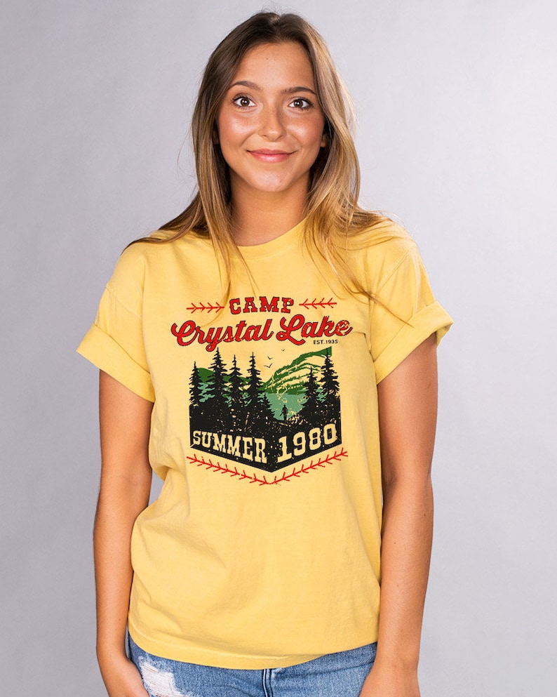May include: Yellow t-shirt with a vintage-style graphic design that says "Camp Crystal Lake Est. 1955 Summer 1980". The graphic features a forest scene with a lake and a person fishing.